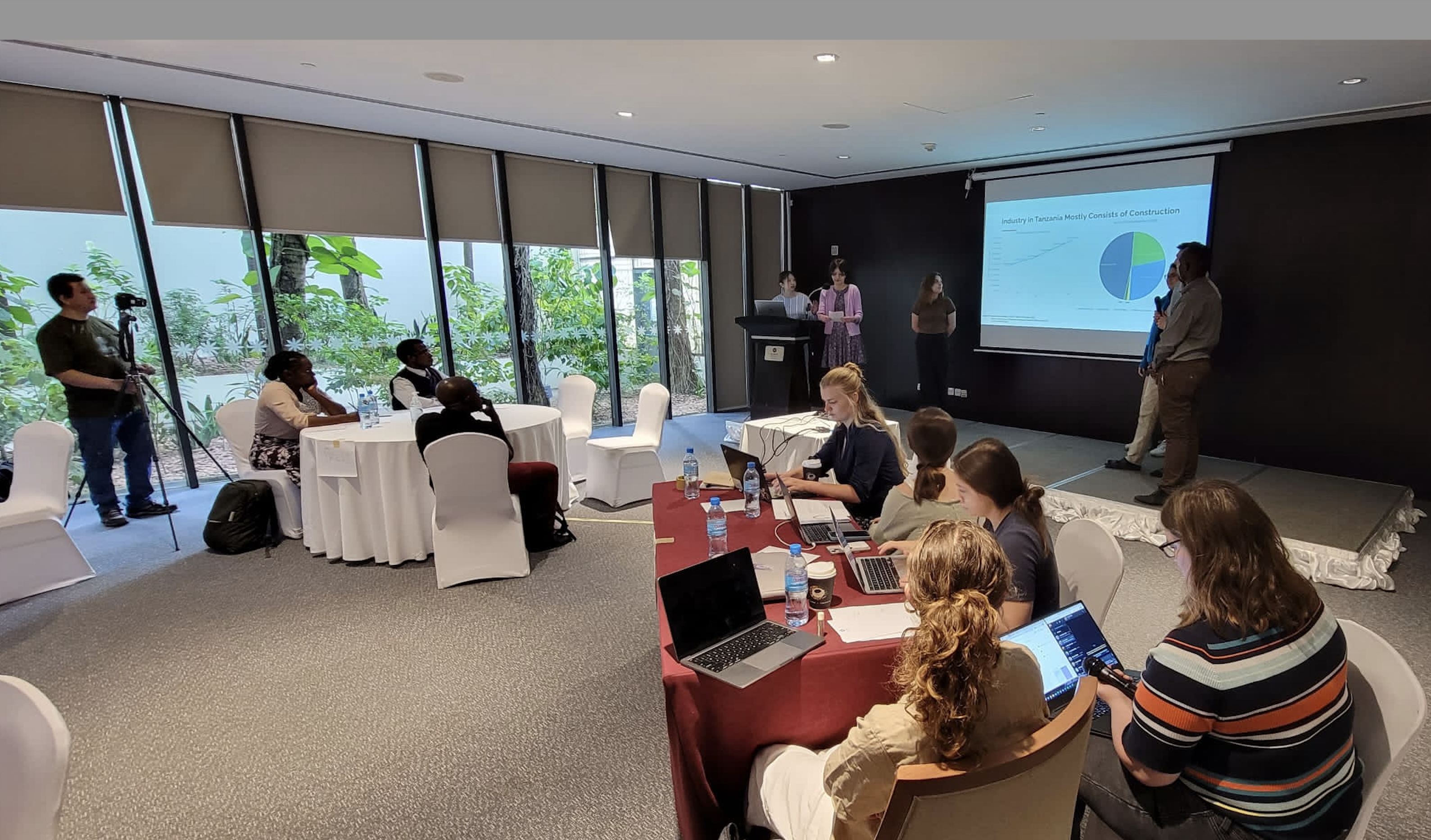 Several students seated at a long table with laptops and notepads, and in the background students presenting about industry in Tanzania.