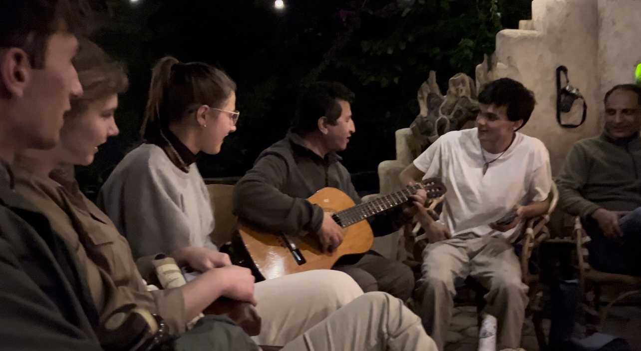 Prasana is playing an acoustic guitar while Rayne, Sophie, and Kaden watch. Off camera there is a fire place near by.